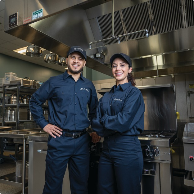 Two smiling Allied Service Group technicians in caps standing in a commercial kitchen
