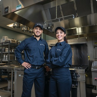 Two smiling Allied Service Group technicians in caps standing in a commercial kitchen