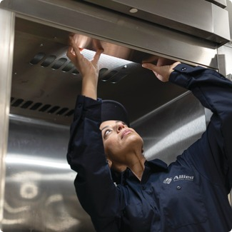 Allied Service Group technician reaching up to inspect stainless steel commercial kitchen equipment