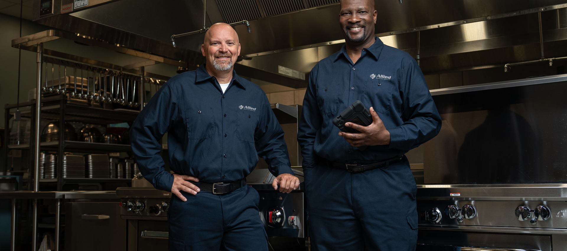 Two Allied Service Group technicians standing in a commercial kitchen, one holding a tablet