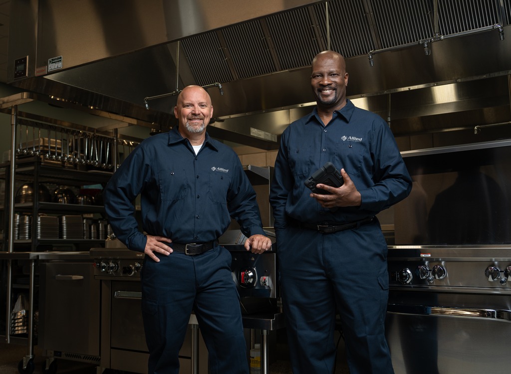 Two Allied Service Group technicians standing in a commercial kitchen, one holding a tablet